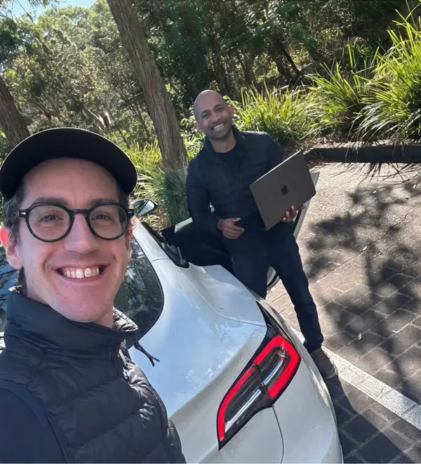 Niklas and Kogulan smiling by a car on a site visit, Kogulan holding a laptop